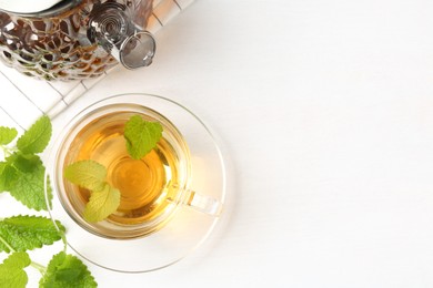 Photo of Aromatic lemon balm tea in glass cup, teapot and fresh leaves on white table, flat lay. Space for text