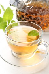 Photo of Aromatic lemon balm tea in glass cup, teapot and fresh leaves on white table, closeup