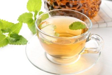 Photo of Aromatic lemon balm tea in glass cup, teapot and fresh leaves on white table, closeup