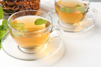 Photo of Aromatic lemon balm tea in glass cups, teapot and fresh leaves on white table, closeup