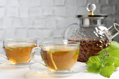 Photo of Aromatic lemon balm tea in glass cups, teapot and fresh leaves on white table, closeup