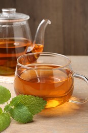 Photo of Aromatic lemon balm tea in glass cup, teapot and fresh leaves on wooden table, closeup