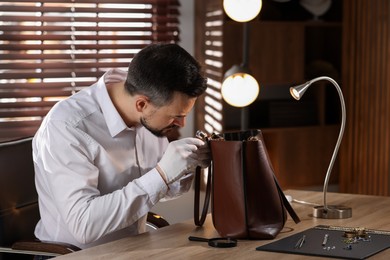 Photo of Appraiser with loupe evaluating leather bag at table indoors