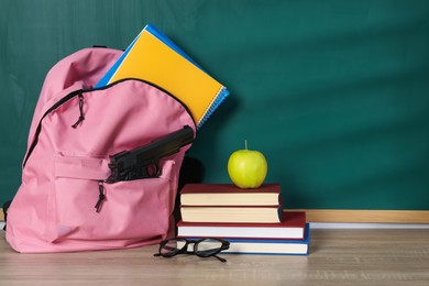Photo of Backpack with gun, stationery, glasses and apple on wooden table near blackboard