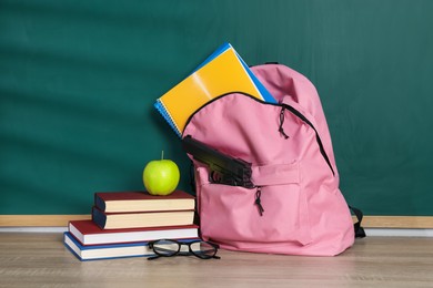 Photo of Backpack with gun, stationery, glasses and apple on wooden table near blackboard