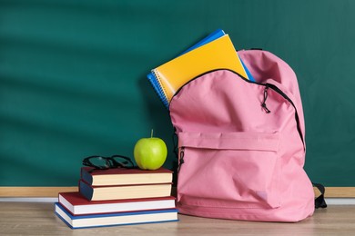 Photo of Backpack with workbooks, books, glasses and apple on wooden table near blackboard
