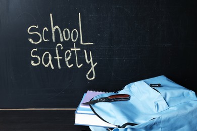 Photo of Backpack with book, notebooks and knife on black wooden table near blackboard. School safety