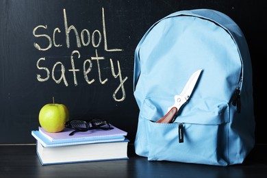 Photo of Backpack with knife, book, notebooks, glasses and apple on black wooden table near blackboard. School safety