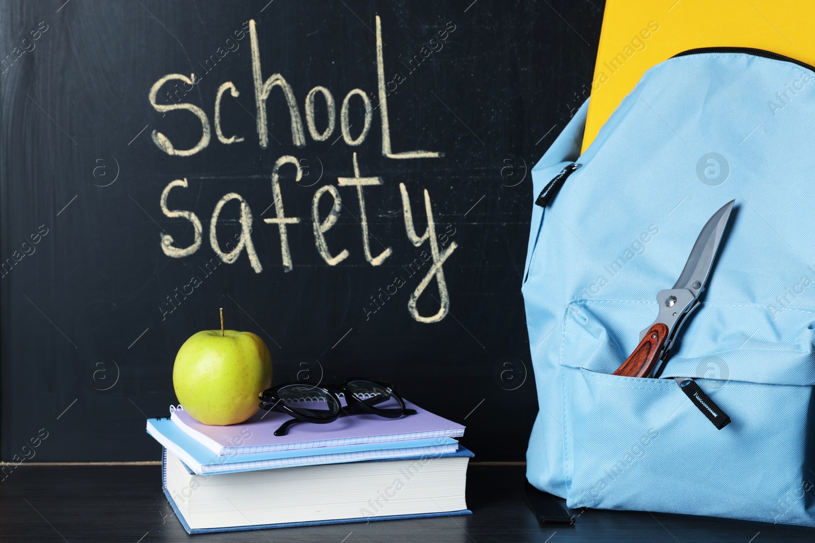 Backpack with knife, book, notebooks, glasses and apple on black wooden table near blackboard. School safety Photo of Backpack with knife, book, notebooks, glasses and apple on black wooden table near blackboard. School safety