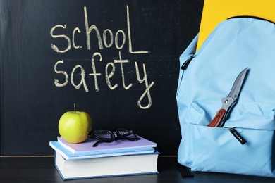 Photo of Backpack with knife, book, notebooks, glasses and apple on black wooden table near blackboard. School safety
