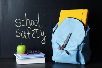 Photo of Backpack with knife, book, notebooks, glasses and apple on black wooden table near blackboard. School safety