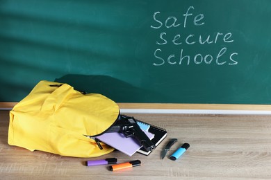 Photo of Backpack, gun, knife and stationery on wooden table near blackboard with words Safe Secure Schools