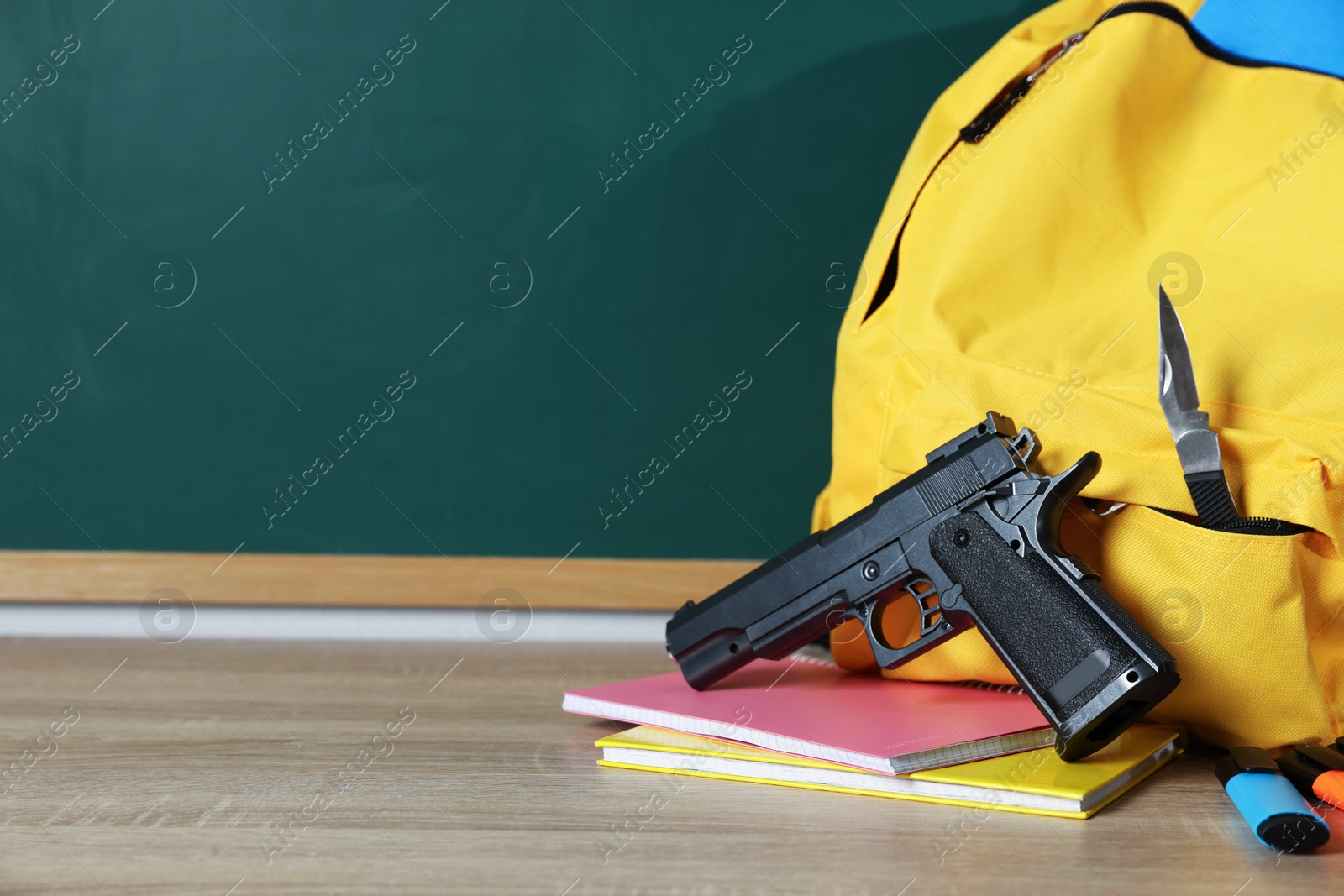 Backpack with knife, gun and stationery on wooden table near blackboard, closeup. Space for text Photo of Backpack with knife, gun and stationery on wooden table near blackboard, closeup. Space for text
