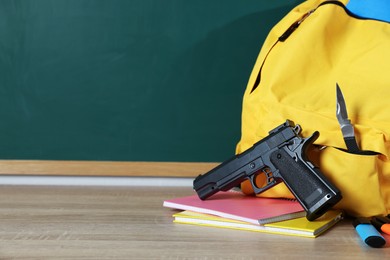 Photo of Backpack with knife, gun and stationery on wooden table near blackboard, closeup. Space for text