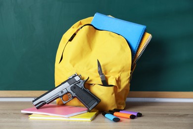 Photo of Backpack with knife, gun and stationery on wooden table near blackboard