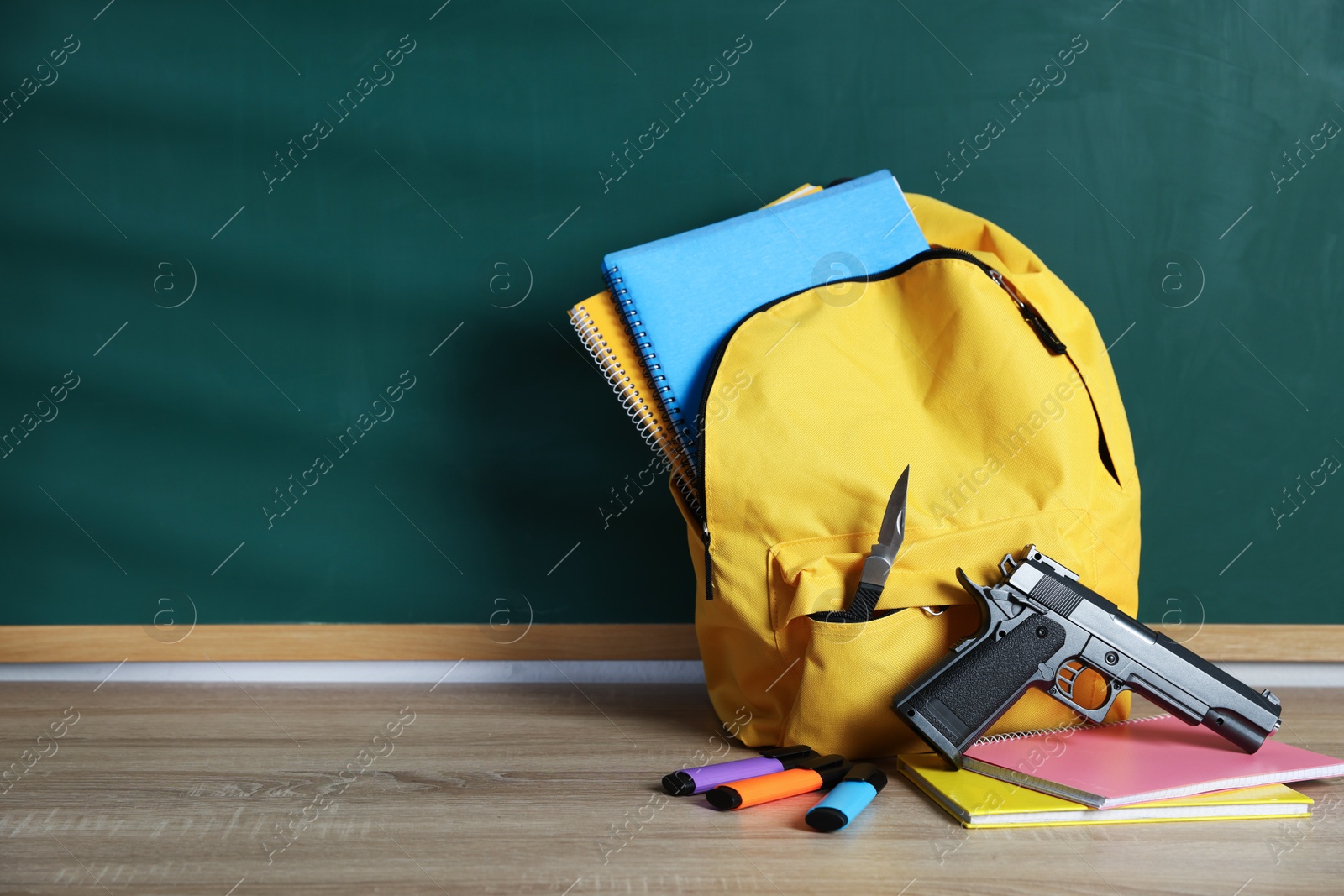 Backpack with knife, gun and stationery on wooden table near blackboard, space for text Photo of Backpack with knife, gun and stationery on wooden table near blackboard, space for text