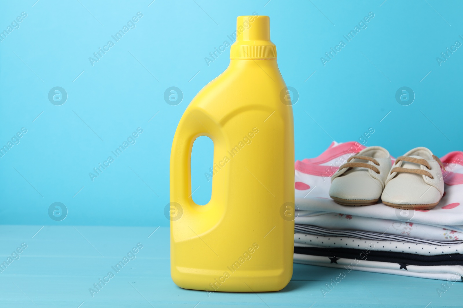 Bottle of fabric softener, stacked baby clothes and shoes on light blue background Photo of Bottle of fabric softener, stacked baby clothes and shoes on light blue background