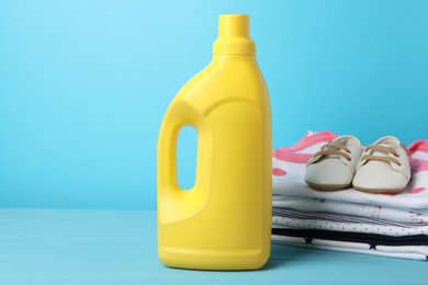 Photo of Bottle of fabric softener, stacked baby clothes and shoes on light blue background