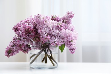 Photo of Beautiful lilac flowers in glass vase on white table indoors, space for text