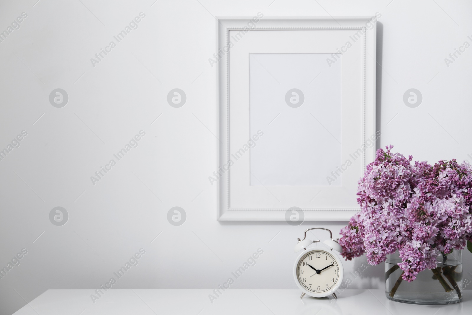 Beautiful lilac flowers and alarm clock on table near white wall indoors, space for text Photo of Beautiful lilac flowers and alarm clock on table near white wall indoors, space for text