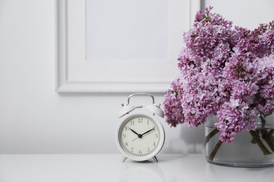Photo of Beautiful lilac flowers and alarm clock on table near white wall indoors, closeup