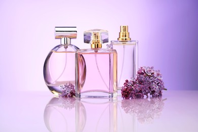 Photo of Bottles of perfume and lilac flowers on mirror surface against violet gradient background