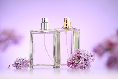 Photo of Bottles of perfume and lilac flowers on mirror surface against violet gradient background, selective focus