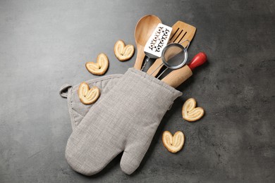 Photo of Different kitchen utensils in oven mitten and cookies on grey table, flat lay