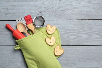 Photo of Different kitchen utensils in oven mitten and palmier cookies on light grey wooden table, flat lay. Space for text