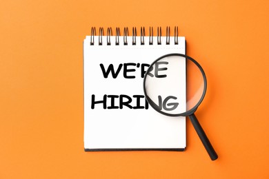 Image of We Are Hiring. Notebook with magnifying glass on orange background, top view. Recruitment, human resources concept