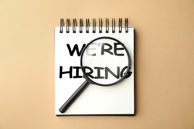 Image of We Are Hiring. Notebook with magnifying glass on beige background, top view. Recruitment, human resources concept