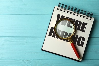 Image of We Are Hiring. Notebook with magnifying glass on light blue wooden table, top view. Recruitment, human resources concept