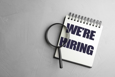 Image of We Are Hiring. Notebook and magnifying glass on grey background, top view. Recruitment, human resources concept
