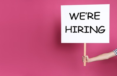 Image of Woman holding sign with phrase We Are Hiring on pink background, closeup. Recruitment, human resources concept
