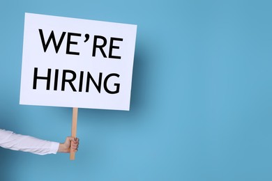 Image of Man holding sign with phrase We Are Hiring on light blue background, closeup. Recruitment, human resources concept