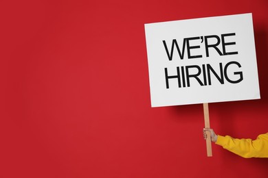 Image of Woman holding sign with phrase We Are Hiring on red background, closeup. Recruitment, human resources concept