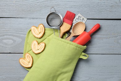 Photo of Different kitchen utensils in oven mitten and palmier cookies on light grey wooden table, flat lay