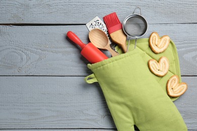 Photo of Different kitchen utensils in oven mitten and palmier cookies on light grey wooden table, flat lay. Space for text