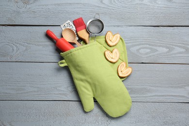 Photo of Different kitchen utensils in oven mitten and palmier cookies on light grey wooden table, flat lay