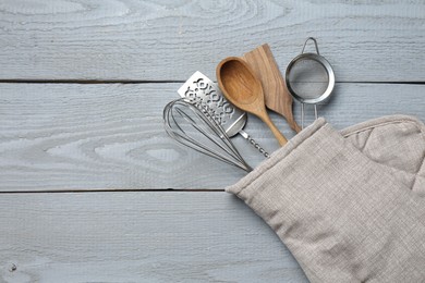 Photo of Different kitchen utensils in oven mitten on light grey wooden table, top view. Space for text