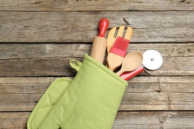 Photo of Different kitchen utensils in oven mitten on wooden table, top view