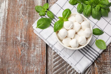 Photo of Tasty mozzarella cheese balls in bowl and basil on wooden table, flat lay. Space for text