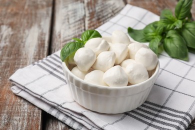 Photo of Tasty mozzarella cheese balls in bowl and basil on wooden table, closeup