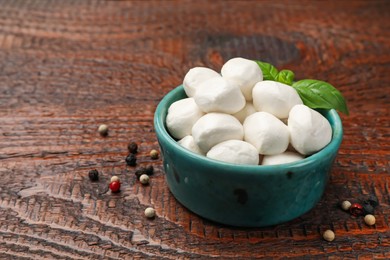 Photo of Tasty mozzarella cheese balls in bowl, peppercorns and basil on wooden table, closeup. Space for text