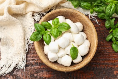 Photo of Tasty mozzarella cheese balls in bowl and basil on wooden table, flat lay