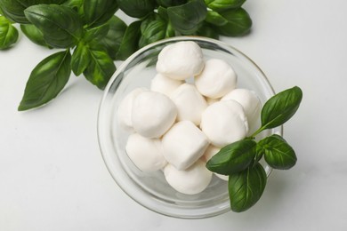 Photo of Tasty mozzarella cheese balls in bowl and basil on white marble table, flat lay
