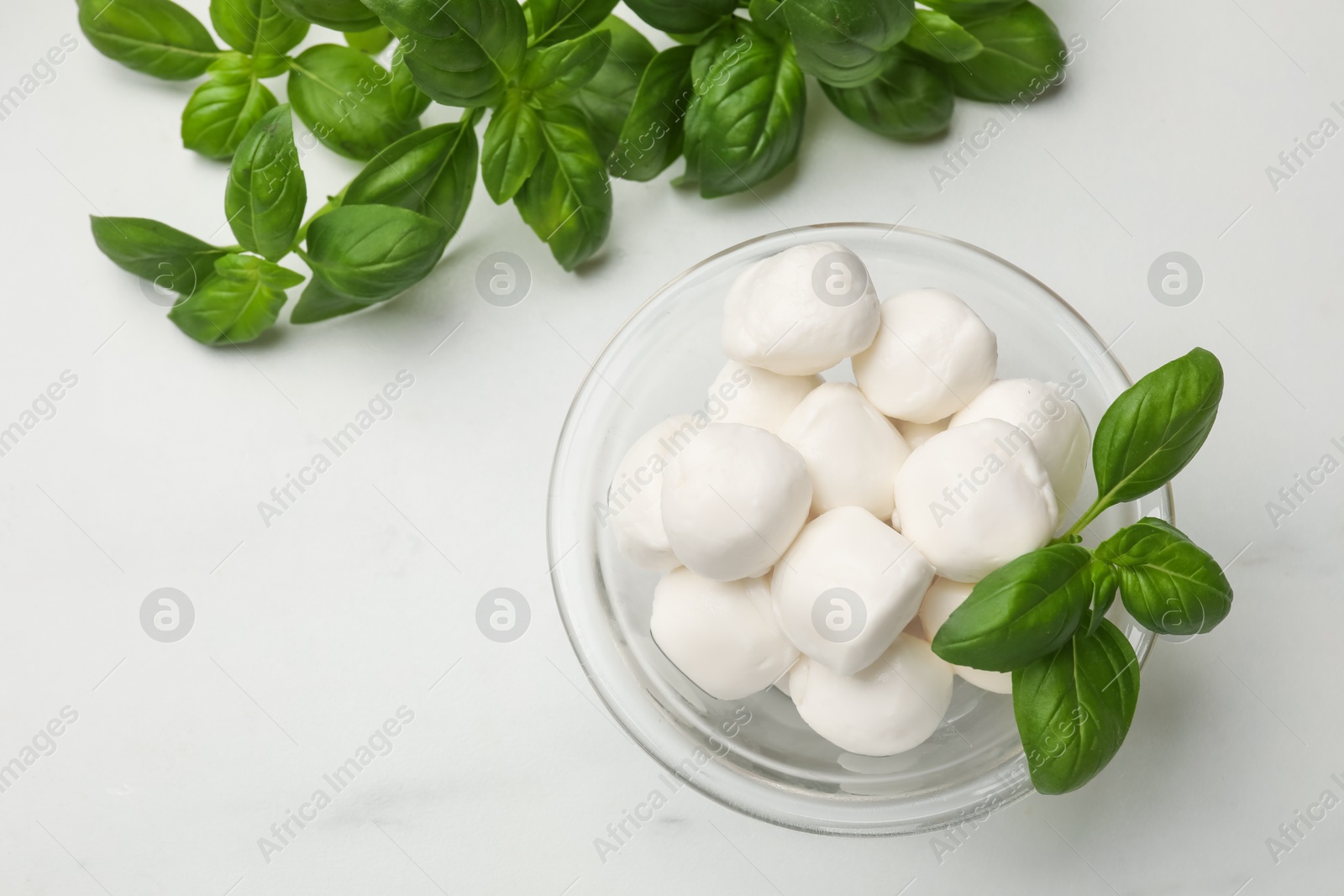 Photo of Tasty mozzarella cheese balls in bowl and basil on white marble table, flat lay. Space for text
