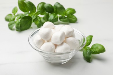 Photo of Tasty mozzarella cheese balls in bowl and basil on white marble table, closeup