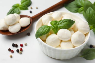 Photo of Tasty mozzarella cheese balls in bowl, spoon, basil and peppercorns on white marble table, closeup
