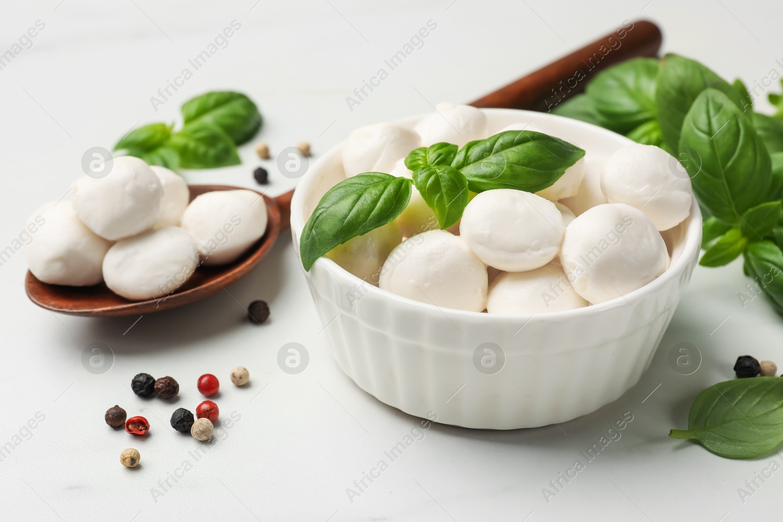 Tasty mozzarella cheese balls in bowl, spoon, basil and peppercorns on white marble table, closeup Photo of Tasty mozzarella cheese balls in bowl, spoon, basil and peppercorns on white marble table, closeup